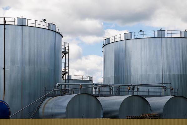 Two large cylindrical metal storage tanks with smaller cylindrical structures and connecting pipes in front, under a partly cloudy sky.